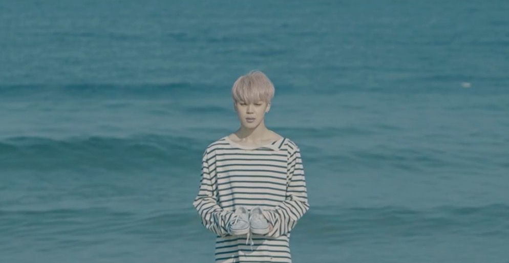 Jimin BTS memegang sepatu di pinggir pantai.