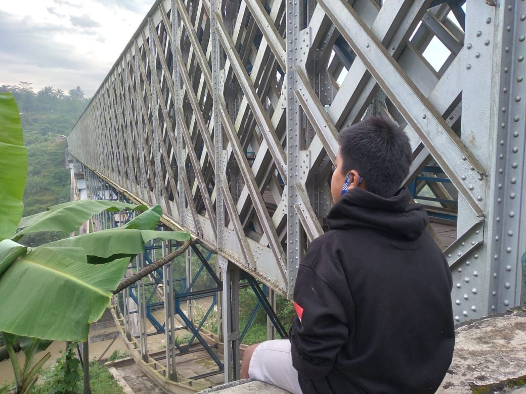 Seorang anak kecil sedang duduk merenung di pinggir jembatan Cirahong yang melintasi sungai Citanduy sementara atasnya jalur kereta api.