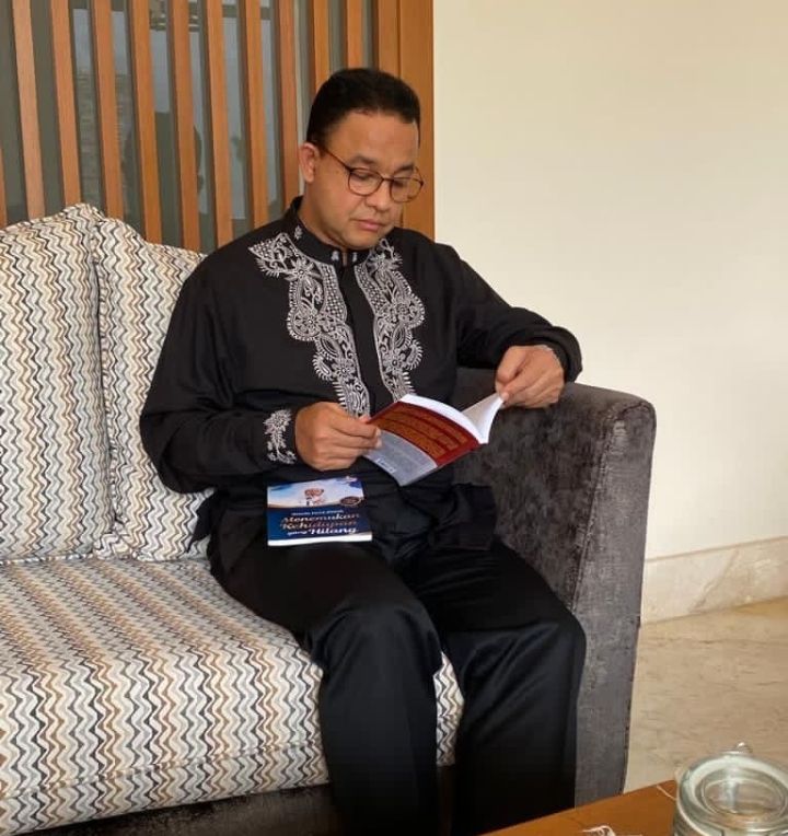 Anies yang mengenakan baju koko berwarna hitam sedang membaca buku karya Ketua Umum Partai Dakwah Rakyat Indonesia (PDRI).