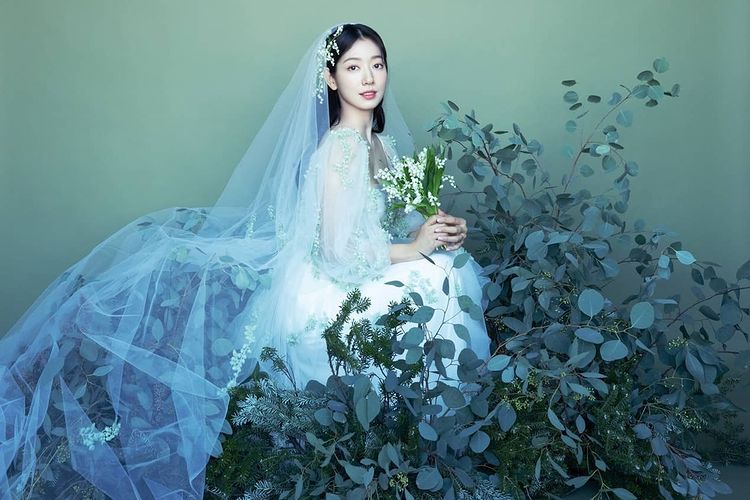 Tampilan foto Park Shin Hye saat kenakan gaun putih dengan veil putih dan mahkota bunga yang dikelilingi dedaunan hijau