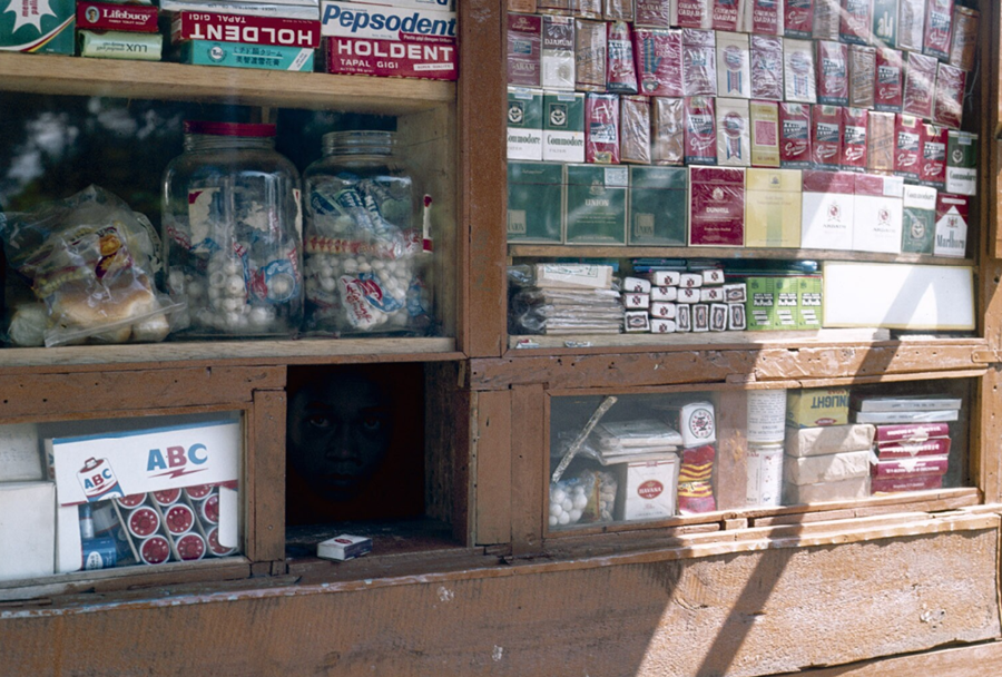 Pemandangan kios rokok di Bandung tahun 1980-an, tampak penjualnya nongol dari lubang kecil.