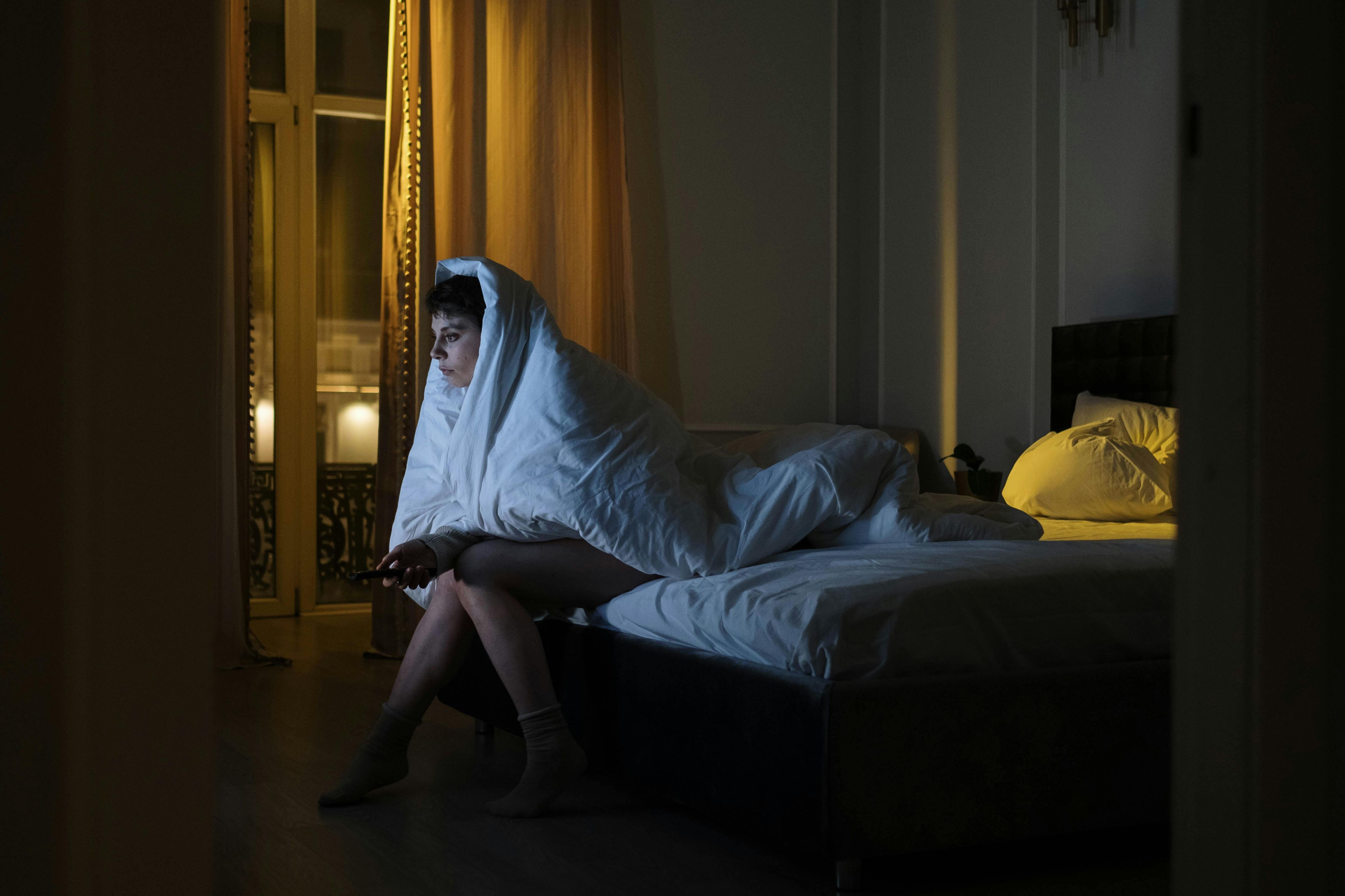 Ilustrai seorang perempuan yang alami gangguan tidur yang sedang menyaksikan siaran televisi di kamar tidurnya.