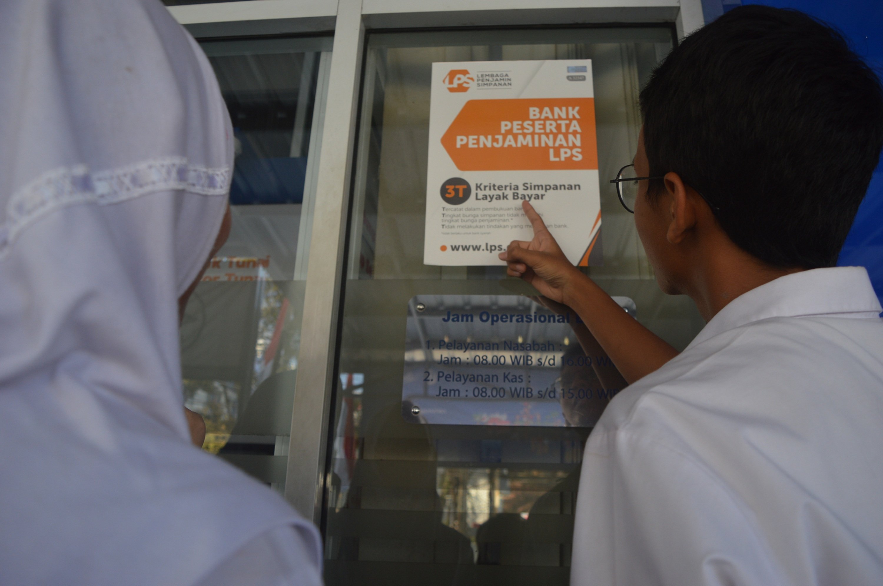 Dua orang pelajar tengah mengamati pamflet LPS yang dipasang di kaca depan kantor BRI Unit Sukarindi